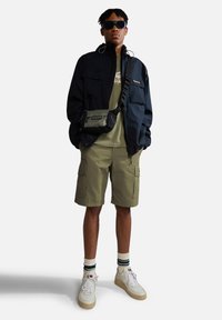 Outfit casual da uomo composto da una giacca nera, una maglietta verde oliva, pantaloni cargo, sneakers bianche e occhiali da sole con una piccola borsa a tracolla.