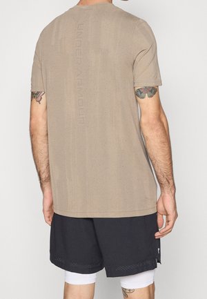 Mężczyzna ubrany w beżową, perforowaną koszulę z krótkim rękawem i czarne szorty, z kolorowymi tatuażami motyli na obu górnych ramionach, widziany od tyłu.