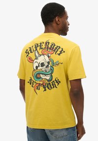 Yellow cotton t-shirt featuring a large graphic print of a skull entwined with snakes and decorative text on the back. Short sleeves, classic fit.