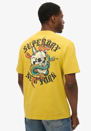 Yellow cotton t-shirt featuring a large graphic print of a skull entwined with snakes and decorative text on the back. Short sleeves, classic fit.