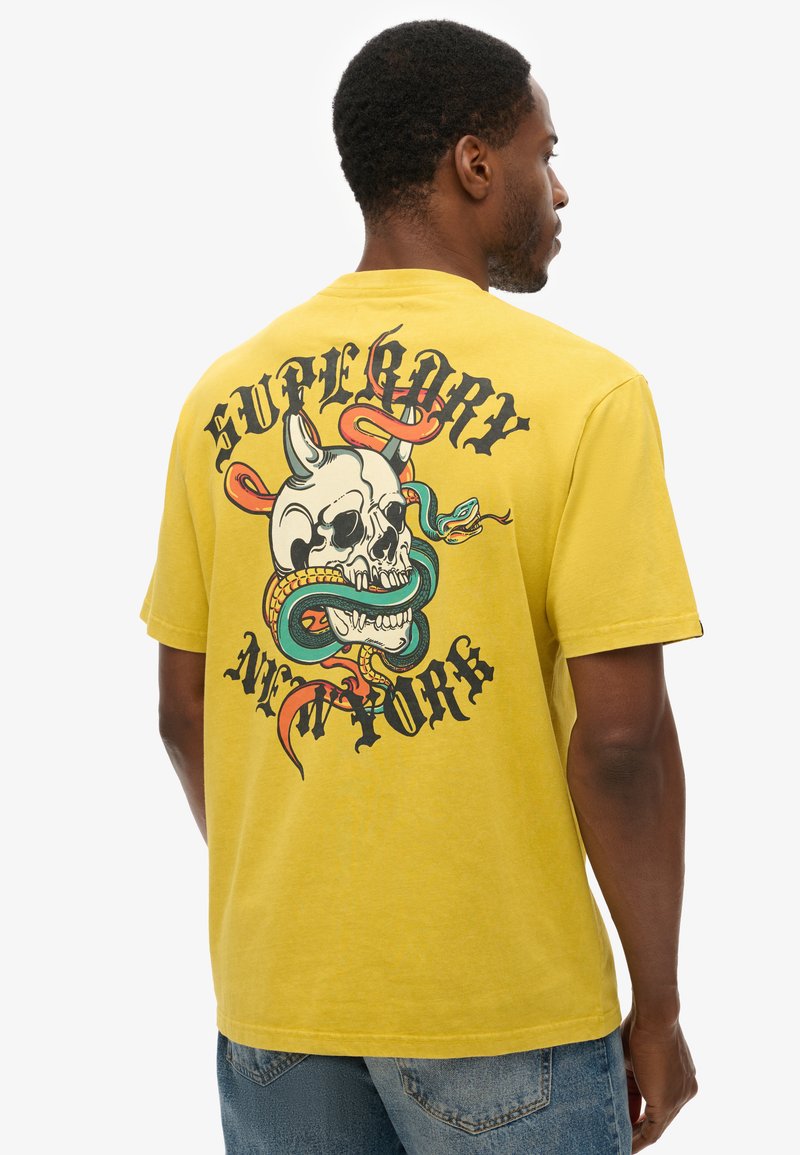 Yellow cotton t-shirt featuring a large graphic print of a skull entwined with snakes and decorative text on the back. Short sleeves, classic fit.