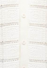 Λευκό πλεκτό ζακέτα με τετράγωνα σχέδια και λεπτομέρειες ανοιχτής πλέξης, με κατακόρυφη σειρά από τέσσερα λευκά κουμπιά που ταιριάζουν.