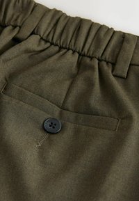 Olivzaļas bikses ar elastīgu jostu, vienu melnu pogu un aizmugurējo kabatu ar gludu auduma faktūru.