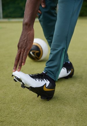 Person in grüner Hose und weißen-schwarz-gelben Fußballschuhen, der im Gras seine Hand zum Fuß streckt, in der Nähe liegt ein Fußball.