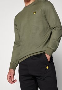 Sudadera de manga larga verde oliva con cuello redondo, con un logo de un pájaro amarillo en el pecho. Combina con pantalones deportivos negros.