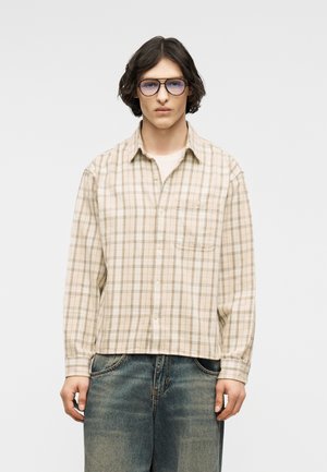 Jeune personne aux cheveux mi-longs et foncés portant des lunettes teintées de bleu, une chemise à carreaux beige boutonnée et un jean bleu délavé, debout devant un fond blanc.