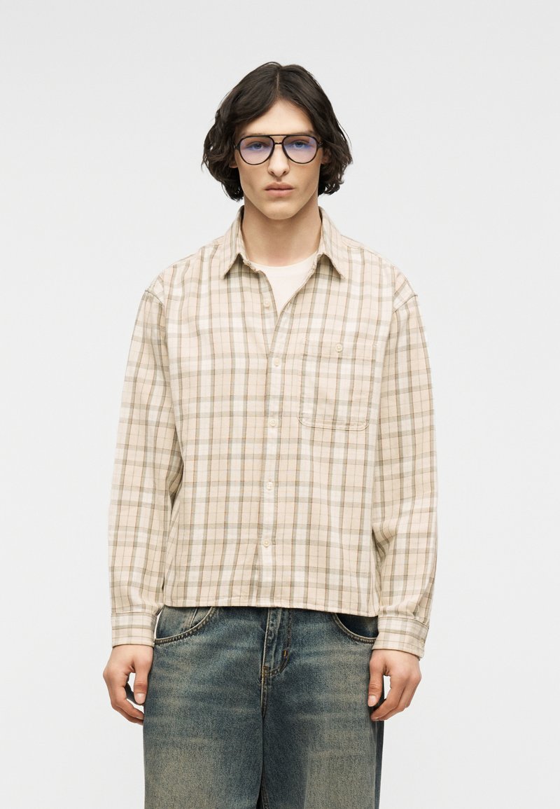 Jeune personne aux cheveux mi-longs et foncés portant des lunettes teintées de bleu, une chemise à carreaux beige boutonnée et un jean bleu délavé, debout devant un fond blanc.