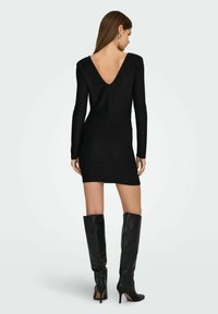 Czarna mini sukienka z długim rękawem, z głębokim dekoltem na plecach w kształcie litery V, gładka w dotyku, dopasowanego kroju, zestawiona z wysokimi czarnymi skórzanymi kozakami.