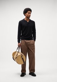 Zwarte lange mouwen polo, bruine wijde broek, met een tan weekendtas met bruine handvatten, zwarte schoenen, staand tegen een lichte achtergrond.