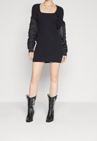 Kvinna iklädd en svart ribbad miniklänning, svart jacka hängande över axlarna och svarta westernstövlar i midthöjd med vit broderi.