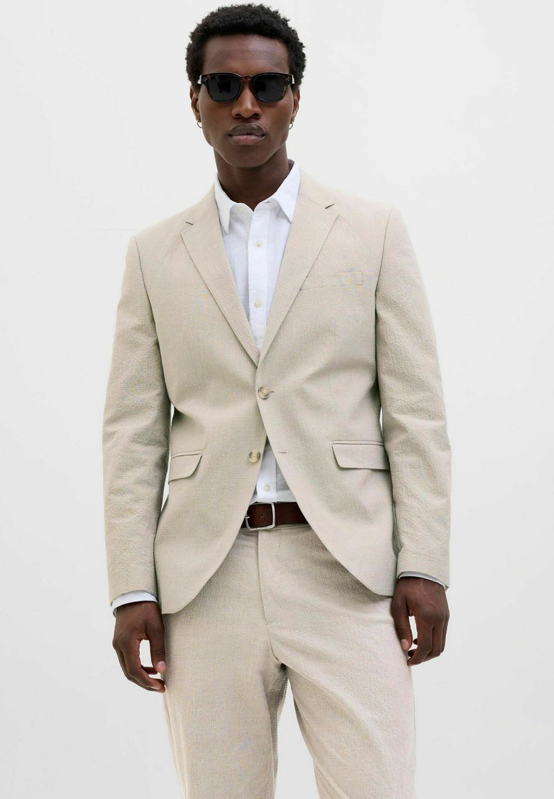 Beige textured suit with a fitted blazer and trousers, paired with a white shirt. Accessorized with sunglasses and a brown belt.