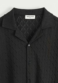 Chemise noire à manches courtes avec un motif en losanges texturé, un col et des fermetures à boutons. Fabriquée en matériau léger. Taille M.