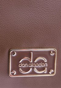 Textura de cuero marrón con una placa rectangular con acentos dorados que muestra "don algodon" y el logo "da". Detalles notables de costura y superficie.