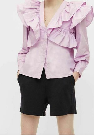 Personne portant un chemisier violet clair à volants avec manches longues et un short noir jusqu'aux genoux, les mains dans les poches.