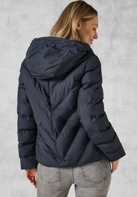 Veste doudoune navy avec capuche, présentant un design en chevrons matelassé et des poignets élastiques. Fabriquée à partir d'un matériau léger et lisse.