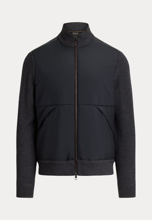 Homme portant une veste zippée noire, un pantalon noir, des chaussures blanches et des lunettes de soleil rondes noires, debout devant un fond beige.