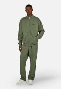 Zaļš sporta krekls un bikses komplekts, izgatavots no mīksta auduma, ar augstu apkakli, rāvējslēdzēju un minimāla logotipa detaļu. Nedaudz atvieglota piegriezuma.