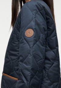 Ναυτικό καπιτονέ μπουφάν με υφή, με καφέ δερμάτινο λογότυπο στο μανίκι και τσέπη με καφέ λεπτομέρεια.