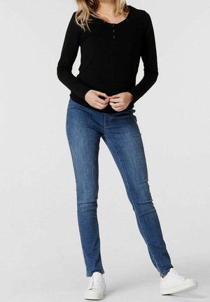 Vrouw draagt een zwarte henley-top met lange mouwen, blauwe skinny jeans en witte sneakers, staat met gevouwen handen voor een effen achtergrond.