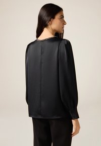 Blusa de satén negra con mangas abullonadas, cuello redondo y adornos de joyas en la parte posterior. Presenta puños abotonados y una textura suave.