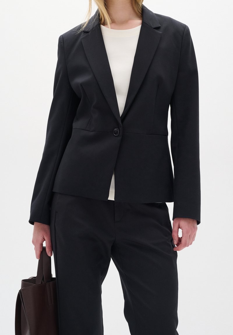 Femme portant un blazer noir sur mesure et un pantalon avec une chemise blanche, tenant un sac fourre-tout marron foncé dans une main sur un fond uni.