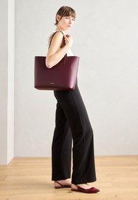 Sac fourre-tout en cuir bordeaux avec une texture lisse, un design vertical et des poignées courtes. Le modèle porte un pantalon large noir et des chaussures bordeaux.