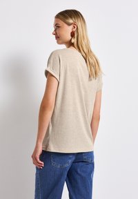 Una camiseta de manga corta beige con un corte relajado, textura suave y puños enrollados, combinada con pantalones de mezclilla azules. Diseño simple y minimalista.
