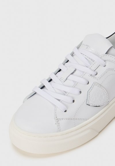Weiße Leder-Sneaker mit flachen weißen Schnürsenkeln, einer strukturierten Gummisohle und einem kleinen Logopatch an der Seite. Glatte Oberfläche mit gestickten Details.
