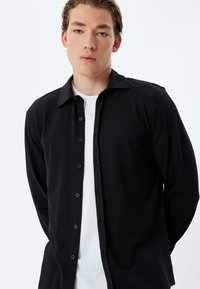 Zwart overhemd met lange mouwen en een kraag, van textuurstof, met knopen aan de voorkant en bijpassende knopen, gedragen over een witte poloshirt.