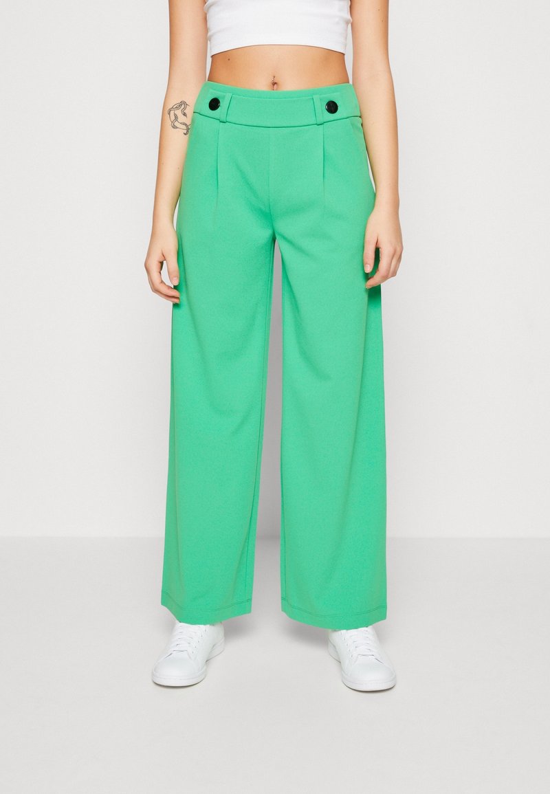 Personne portant un pantalon vert taille haute à jambes larges avec des boutons noirs et des baskets blanches, un haut court blanc, un tatouage sur le bras gauche.