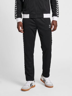 Hombre con pantalones deportivos negros con franjas en chevron blancas en los laterales y zapatillas blancas con detalles negros y suelas de goma.