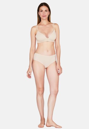 Kvinde stående barfodet iført beige bh og matchende undertøj mod en ensfarvet hvid baggrund.