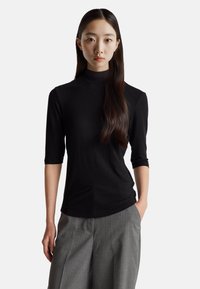 Černý žebrovaný rolák s tříčtvrtečními rukávy, který se vyznačuje těsným střihem a hladkou texturou, spárovaný se šedými plisovanými kalhotami.