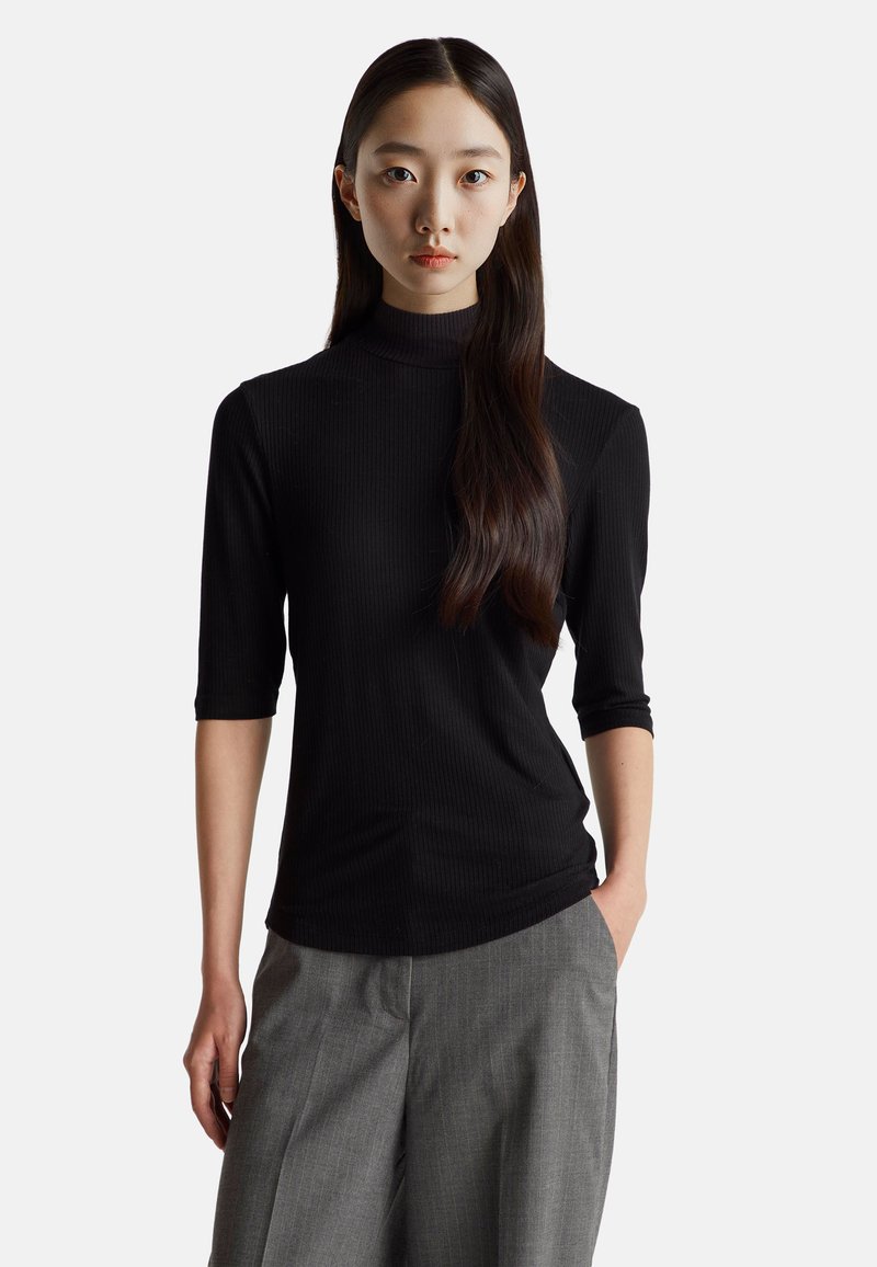 Černý žebrovaný rolák s tříčtvrtečními rukávy, který se vyznačuje těsným střihem a hladkou texturou, spárovaný se šedými plisovanými kalhotami.