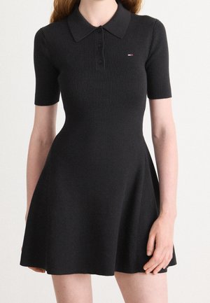 Donna che indossa un abito polo nero aderente in maglia a coste con maniche corte e gonna svasata, con un piccolo logo sul petto.