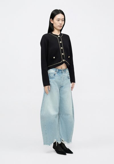 Fekete, rövid szabású kardigán aranylánc díszítéssel és nagy gombokkal, lazán viselt, világoskék, széles szárú, szakadt farmerral és fekete bokacsizmával párosítva.