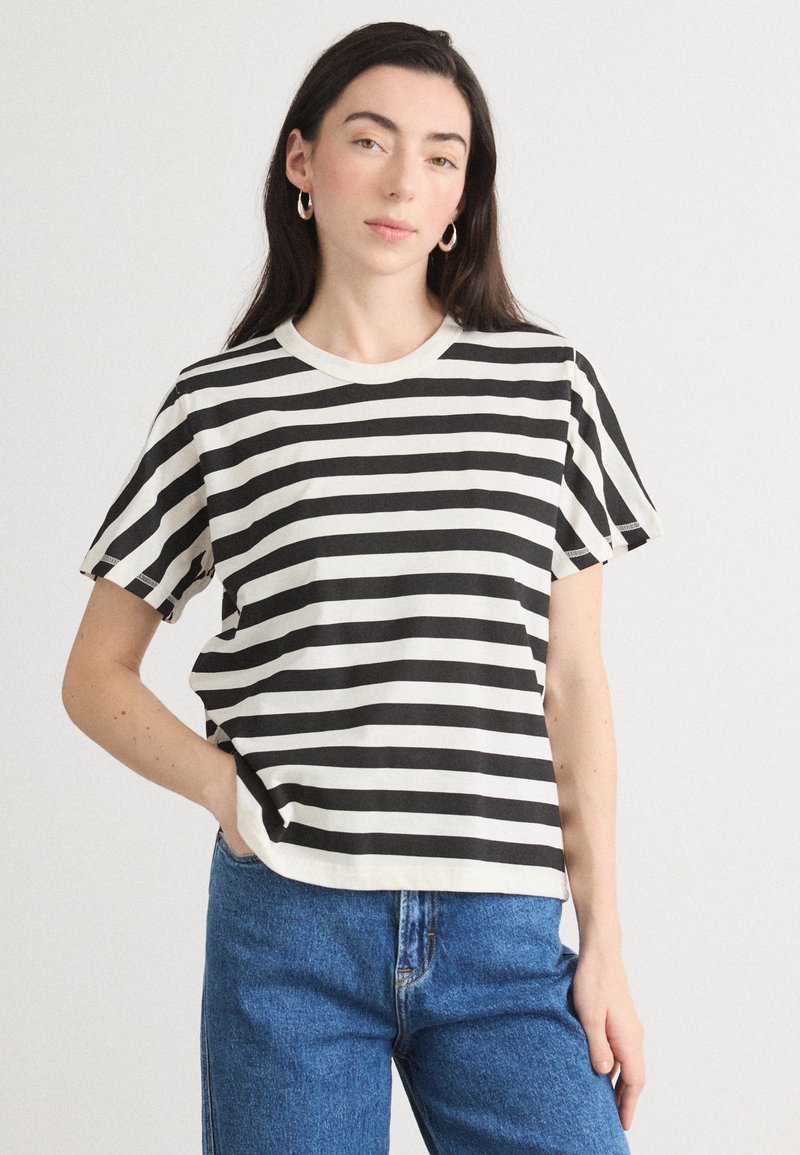 Tricou cu dungi alb-negru, cu decolteu rotund și mâneci scurte, fabricat din bumbac moale, purtat cu blugi blue cu talie înaltă.