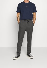 Man in een marineblauwe t-shirt met klein logo en grijze geruite broek met trekkoord, met zwart-witte sneakers, staand met zijn handen in de zakken.