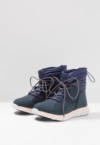 Zapatos deportivos de media altura en tonos verde azulado y marino, con un cuello acolchado, cordones grises y una suela de color melocotón claro. Presentan una parte superior de malla para mayor transpirabilidad.