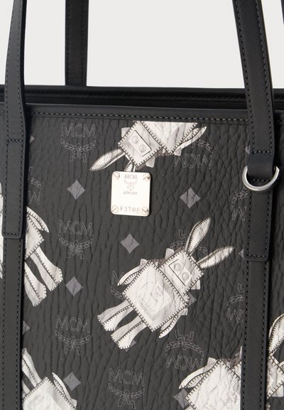 Μαύρη τσάντα tote από δέρμα με ραμμένα λευκά σχέδια ρομπότ και διαμαντιών, ασημένια πλακέτα με την επωνυμία "MCM München" και σειριακό αριθμό F3708.