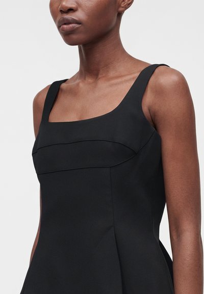 Γυναίκα με φόρεμα χωρίς μανίκια, μαύρο, με τετράγωνη λαιμόκοψη και εφαρμοστό μπούστο σε απλό ανοιχτό φόντο.