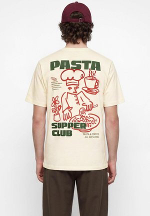 Personne portant un T-shirt crème avec un graphisme de chef bordeaux, assiette de pâtes, tasse de café et texte disant "Pasta Supper Club" et "Pasta & Coffee All Day Long."