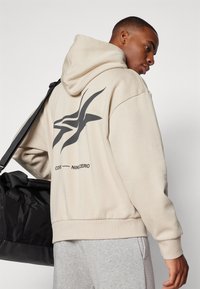 Felpa beige con grafiche nere sulla schiena, caratterizzata da una tasca a marsupio e polsini a coste. Il modello porta una borsa da palestra nera.