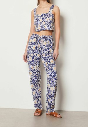 Kobieta nosząca niebiesko-biały top w kwiatowy wzór i dopasowane, wysokie spodnie wraz z brązowymi, otwartymi sandałami na płaskim obcasie.