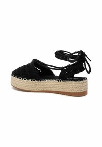 Sandal con cuña de yute tejida en negro con lazos, plataforma de yute y parte superior texturizada. Cuenta con una suela exterior plana de goma marrón.