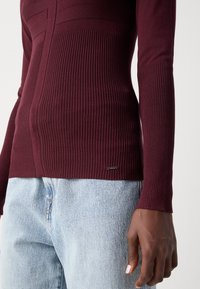 Haut en tricot côtelé bordeaux à manches longues, comportant des lignes horizontales et une petite étiquette de logo. Assorti à un jean en denim bleu clair.