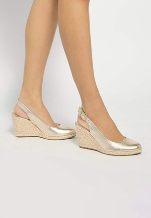 Piernas con zapatos de cuña slingback dorados metálicos con suelas de cuerda trenzada y correas con hebilla sobre un fondo neutro.