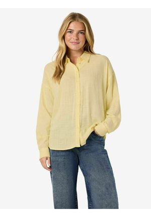 Žlutá lehká košile na knoflíky s límečkem a dlouhými rukávy, kombinovaná s modrými džínami. Látka má měkký, texturovaný vzhled.