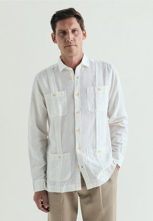 Hombre con cabello corto y oscuro que lleva una camisa blanca abotonada con cuatro bolsillos delanteros y pantalones beige, de pie frente a un fondo neutro.