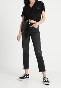 Camicia nera accorciata con bottoni e design in occhiello, annodata in vita; jeans neri strappati con perline e orlo sfrangiato; sneakers nere.